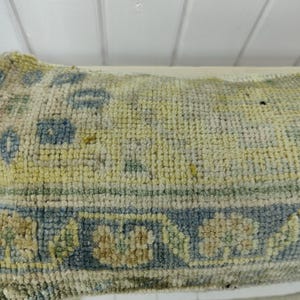 May include: A close-up of a light blue and cream colored rug with a geometric pattern. The rug has a worn and faded appearance.