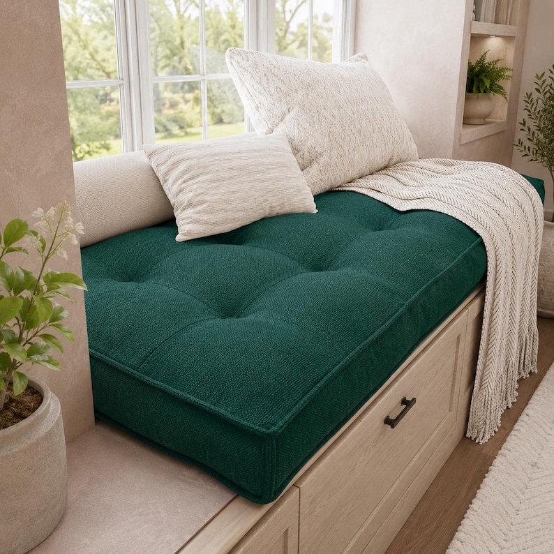 May include: A deep emerald green tufted bench cushion with a textured fabric surface. The cushion is topped with two off-white throw pillows and a cream-colored knit throw blanket with fringe, resting on a light wood storage unit with drawers.