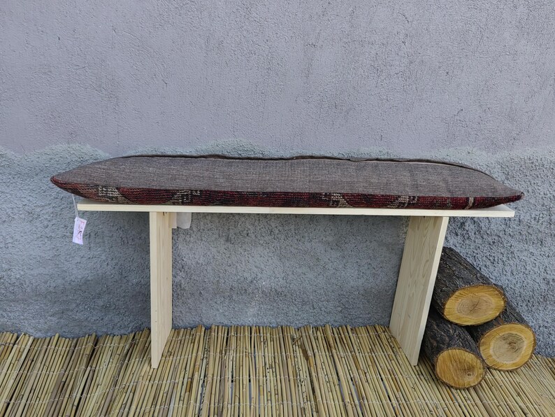 May include: A wooden bench with a patterned cushion. The bench has two legs on each side and is sitting on a woven bamboo mat.