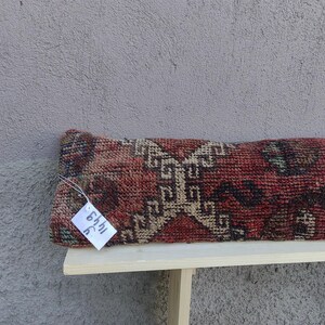 May include: A long, rectangular pillow with a red, brown, and beige patterned fabric. The pillow has a tag attached to it that says "54hl 3C".