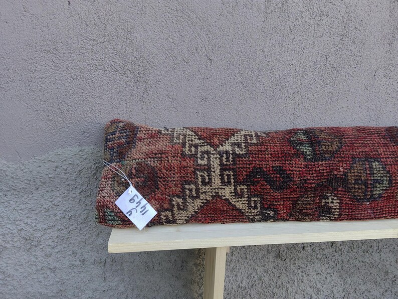 May include: A long, rectangular pillow with a red, brown, and beige patterned fabric. The pillow has a tag attached to it that says "54hl 3C".