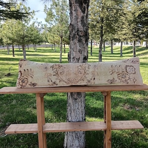 Puede incluir: Un cojín rectangular beige con patrones florales marrones descansa sobre una estantería de madera. El diseño del cojín presenta motivos florales y de volutas simétricos. La estantería forma parte de una estructura de madera, sobre un fondo de árboles y hierba verde.