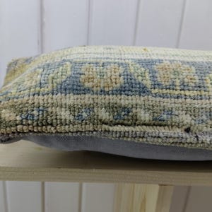 May include: A rectangular pillow with a blue, beige, and green patterned fabric. The pillow has a light gray backing and is sitting on a wooden surface.