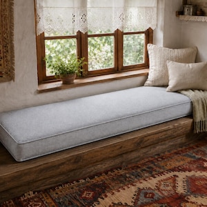 May include: A long, rectangular light grey bench cushion with piped edges, positioned on a wooden window seat. The cushion is accompanied by two beige throw pillows and a draped beige knit throw blanket, creating a soft seating arrangement.