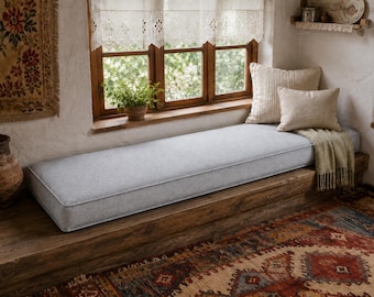 May include: A long, rectangular light gray bench cushion with piped edges, positioned on a wooden window seat. The cushion is accompanied by two beige throw pillows and a draped beige knit throw blanket, creating a soft seating arrangement.