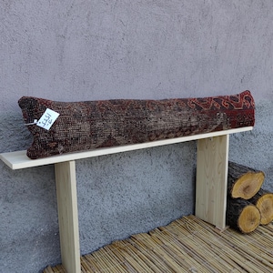 May include: A wooden bench with a long, rectangular cushion covered in a colourful, patterned fabric. The cushion has a white tag with the word "الذكرى" written on it. The bench is sitting on a woven mat.