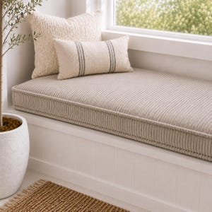 May include: A beige corduroy bench cushion sits on a white window seat. Behind it are two pillows: one square, textured cream pillow and one rectangular cream pillow featuring two vertical gray stripes. The cushion has piped edges.