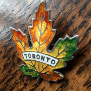 May include: A silver-toned enamel pin in the shape of a maple leaf with the word "TORONTO" in white lettering on a yellow banner. The leaf is orange, yellow, and green.