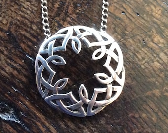 Op de afbeelding: Een zilveren ketting met een ronde hanger in de vorm van een Keltische knoop met ingewikkelde, verweven patronen. De hanger hangt aan een zilveren ketting.