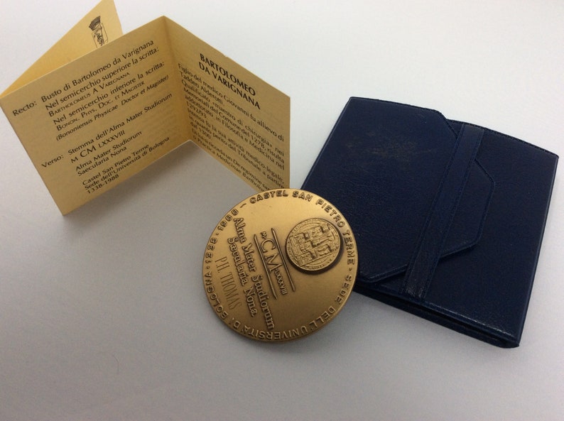 K&ouml;nnte beinhalten: Eine goldfarbene Medaille mit der Inschrift "Alma Mater Studiorum Secularia Nona" und dem Text "CM PIL THOMAS" auf der Vorderseite. Die R&uuml;ckseite der Medaille zeigt ein Wappen mit dem Text "CASTEL SAN PIETRO TERME - 1338 - 1988" und dem Text "UNIVERSITA' DEGLI STUDI DI BOLOGNA" in einem Kreis um das Wappen.