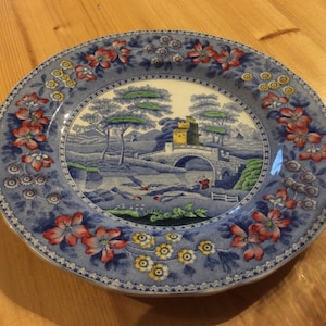 May include: A blue and white ceramic plate with a floral border and a central image of a bridge over a stream with a small building in the background.