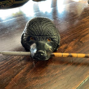 Puede incluir: Un recipiente negro con forma de perro con ojos naranjas, que sostiene un bolígrafo de plata y madera. El recipiente tiene una superficie texturizada. El bolígrafo es plateado con un agarre texturizado y un eje de madera con un patrón marrón y negro.