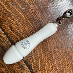 May include: A white ceramic bottle stopper with a twisted metal handle. The stopper has a rounded handle and a conical tip. The text "ROYAL DOULTON" is printed on the handle in a circular emblem.