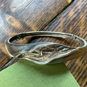 May include: A silver bangle bracelet with a textured, organic design. The bracelet is open and has a hinged clasp.