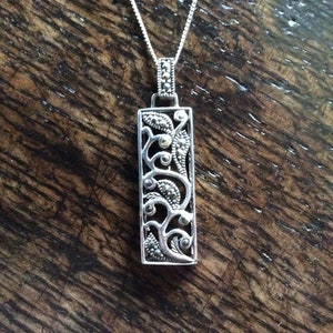 Op de afbeelding: Een zilveren ketting met een rechthoekige hanger met een delicaat bloemenpatroon met marcasietstenen.