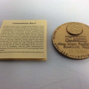 K&ouml;nnte beinhalten: Eine goldfarbene Medaille mit dem Text "Alma Mater Studiorum Sacularia Nona" und "PH. THOMAS" darauf. Die Medaille ist von einem dekorativen Rand umgeben.
