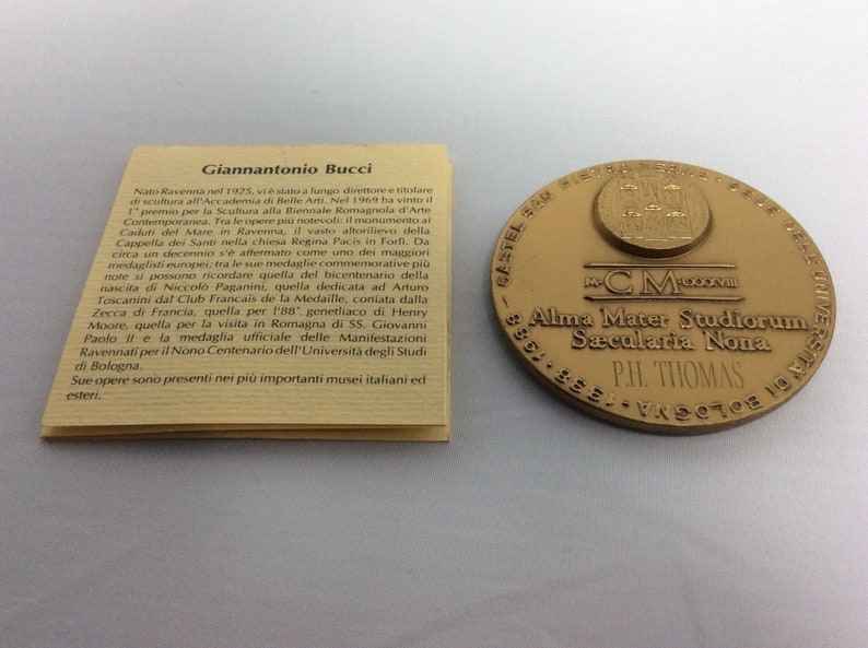 K&ouml;nnte beinhalten: Eine goldfarbene Medaille mit dem Text "Alma Mater Studiorum Sacularia Nona" und "PH. THOMAS" darauf. Die Medaille ist von einem dekorativen Rand umgeben.