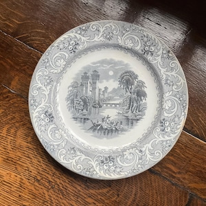 May include: A round ceramic plate featuring a detailed grey and white transferware pattern. The centre depicts a landscape with a castle, a bridge, and a boat on water. The wide rim is decorated with an intricate floral and scrollwork border design.