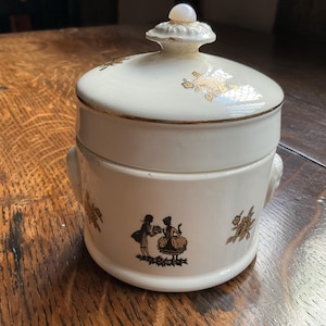 May include: White ceramic jar with a lid and gold accents. The jar features a silhouette of two people in a romantic pose and floral designs.