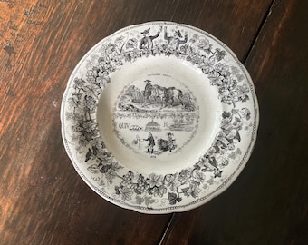 Peut inclure: Assiette ancienne en céramique blanche avec une illustration en noir et blanc d'une scène rurale. L'assiette présente une bordure décorative de vignes et de feuilles. Le centre de l'assiette comporte du texte et un dessin détaillé de personnes et d'animaux.
