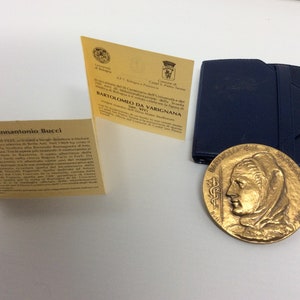 K&ouml;nnte beinhalten: Eine goldfarbene Medaille mit einem Frauenportr&auml;t auf der einen Seite und Text auf der anderen. Die Medaille befindet sich in einem blauen Etui mit einer gelben Karte, auf der "Bartolomeo da Varignana 1567-1643" steht.