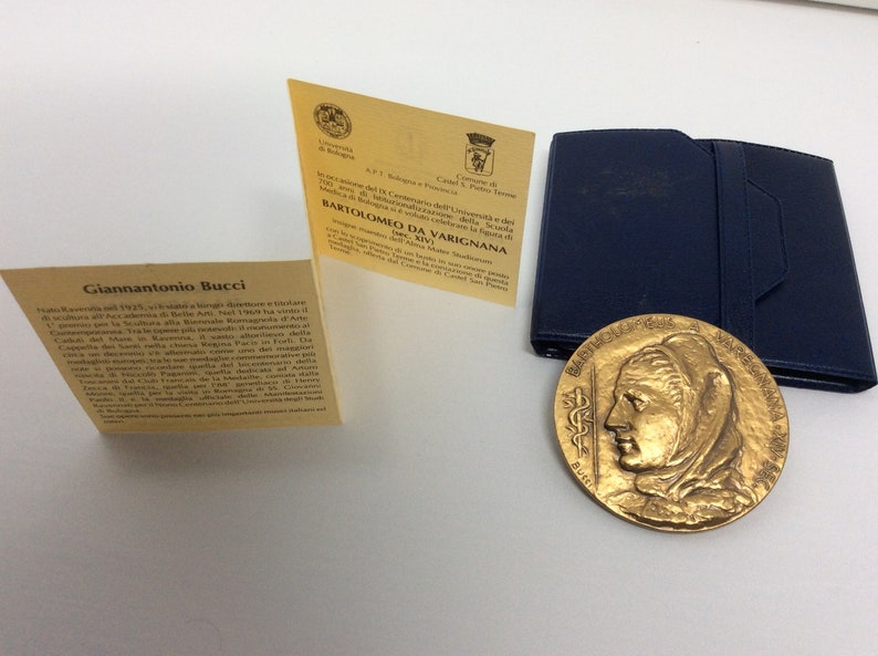 K&ouml;nnte beinhalten: Eine goldfarbene Medaille mit einem Frauenportr&auml;t auf der einen Seite und Text auf der anderen. Die Medaille befindet sich in einem blauen Etui mit einer gelben Karte, auf der "Bartolomeo da Varignana 1567-1643" steht.