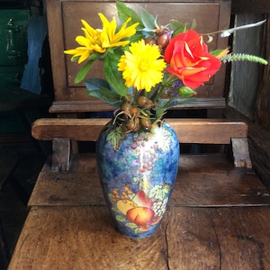May include: A vibrant floral display in a patterned vase. The vase features a blue background with fruit and grape motifs. The arrangement includes yellow and red flowers with green leaves. The vase rests on a wooden surface.
