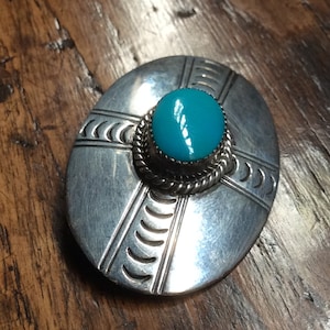 Puede incluir: Un broche ovalado de plata con una piedra turquesa en el centro. El broche tiene un diseño geométrico con un patrón de cruz y un borde de cuerda alrededor de la piedra.