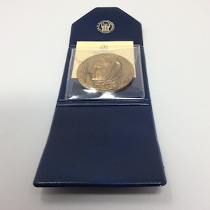 K&ouml;nnte beinhalten: Eine goldfarbene Medaille in einem blauen Lederetui mit einer durchsichtigen Plastikabdeckung. Die Medaille zeigt das Profil einer Person mit einer Krone.