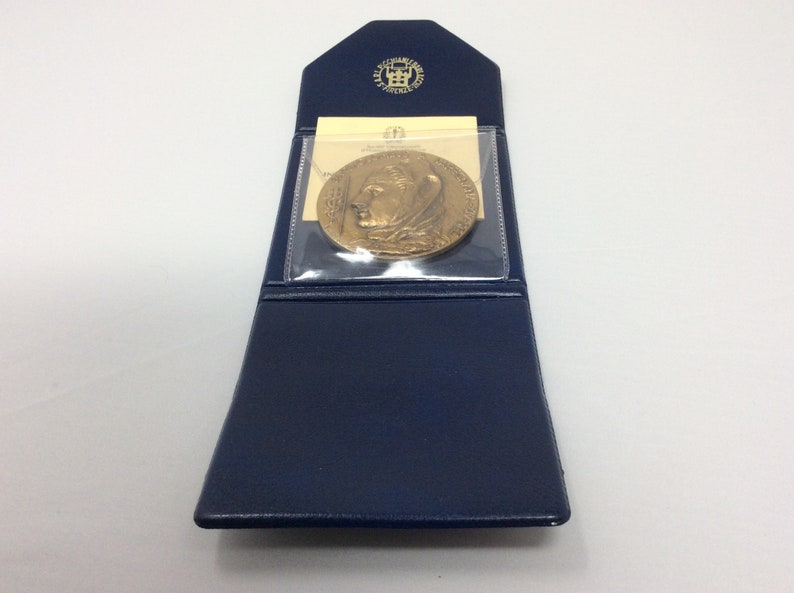 K&ouml;nnte beinhalten: Eine goldfarbene Medaille in einem blauen Lederetui mit einer durchsichtigen Plastikabdeckung. Die Medaille zeigt das Profil einer Person mit einer Krone.