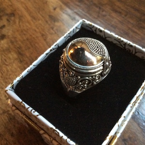May include: A silver ring with a hinged top, featuring an ornate design and a gold-toned interior. The ring is displayed in a white box with a black velvet lining.