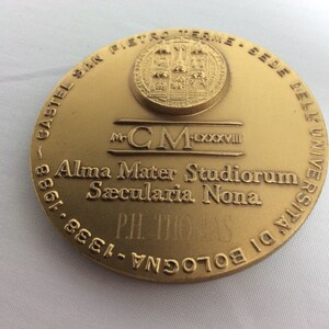 K&ouml;nnte beinhalten: Eine goldfarbene Medaille mit der Inschrift "CASTEL SAN PIETRO TERME - SEDE DELL'UNIVERSITA DI BOLOGNA - 1338 - 1988" am Rand. In der Mitte der Medaille befindet sich ein Wappen mit dem Text "M-CM-LXXXVIII Alma Mater Studiorum S&aelig;cularia Nona PH. THOMAS".
