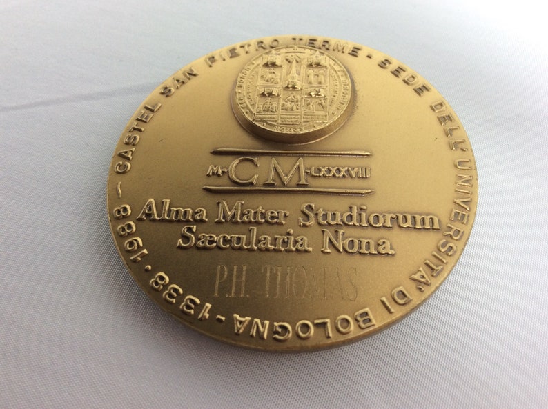 K&ouml;nnte beinhalten: Eine goldfarbene Medaille mit der Inschrift "CASTEL SAN PIETRO TERME - SEDE DELL'UNIVERSITA DI BOLOGNA - 1338 - 1988" am Rand. In der Mitte der Medaille befindet sich ein Wappen mit dem Text "M-CM-LXXXVIII Alma Mater Studiorum S&aelig;cularia Nona PH. THOMAS".