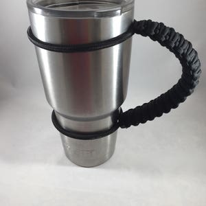 May include: A stainless steel tumbler with a black paracord handle. The tumbler has a lid and the word "YETI" is visible on the side.
