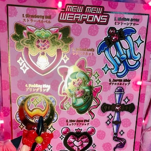 Op de afbeelding: Een roze animefiguur met een bijpassende roze achtergrond met "Mew Mew Weapons" en Japanse tekst. Het figuur houdt een toverstaf vast. De achtergrond heeft illustraties van verschillende wapens.