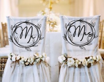 Puede incluir: Dos sillas blancas con guirnaldas de flores blancas y fundas de silla de tela transparente. Las sillas están decoradas con letreros en blanco y negro que dicen "Mr." y "Mrs." en un diseño de corona.