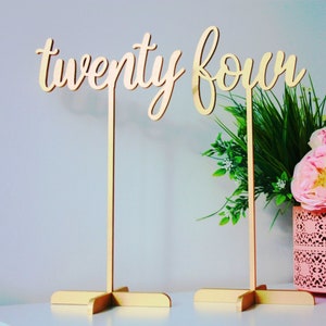 May include: Two gold wooden table numbers, one with the word "twenty" and the other with the word "four", on stands with a cross base. The numbers are in front of a pink vase with green and pink flowers.