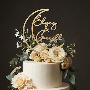 Puede incluir: Un pastel blanco de tres niveles decorado con rosas color crema, flores de jazmín blancas y hojas de eucalipto. Un adorno dorado para pastel en forma de media luna presenta el texto en cursiva Stacy & Garrett centrado en la luna.