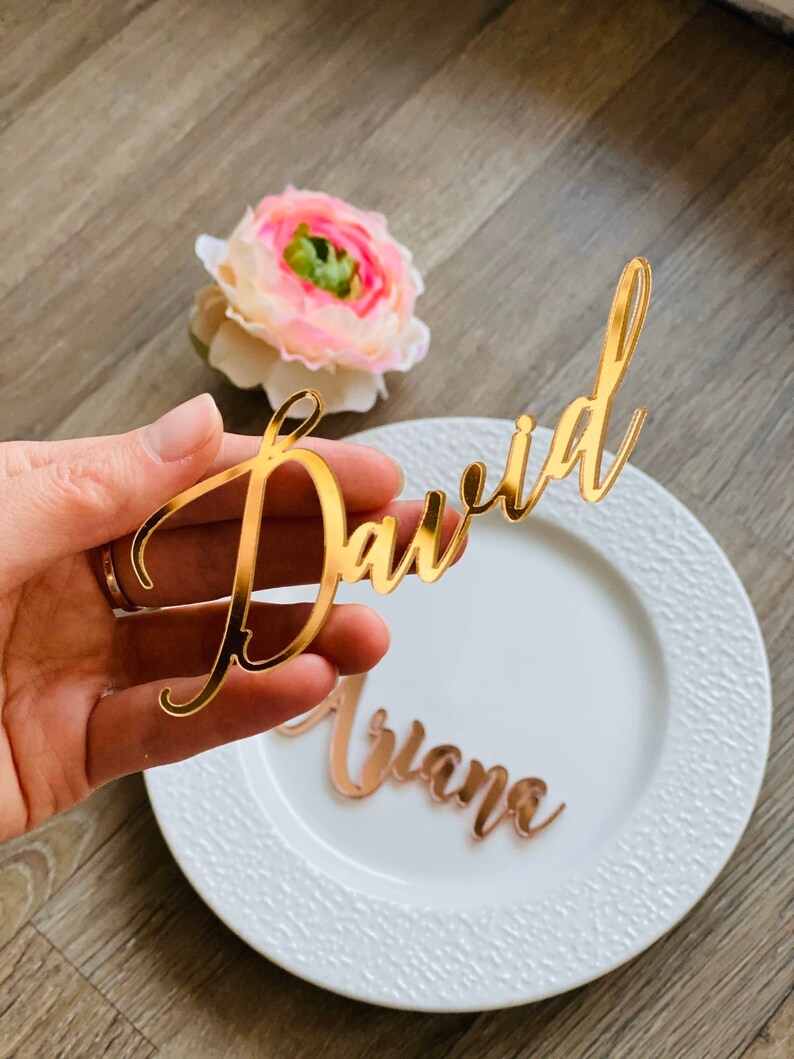 Puede incluir: Tarjetas de lugar de acr&iacute;lico dorado con los nombres "David" y "Ariana" escritos en letra cursiva.