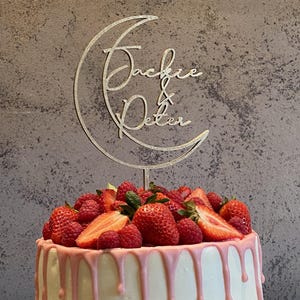 Op de afbeelding: Een witte cake met roze glazuur en verse aardbeien en frambozen. Een houten caketopper met een halve maan ontwerp en de namen "Jackie & Peter" is op de cake geplaatst.
