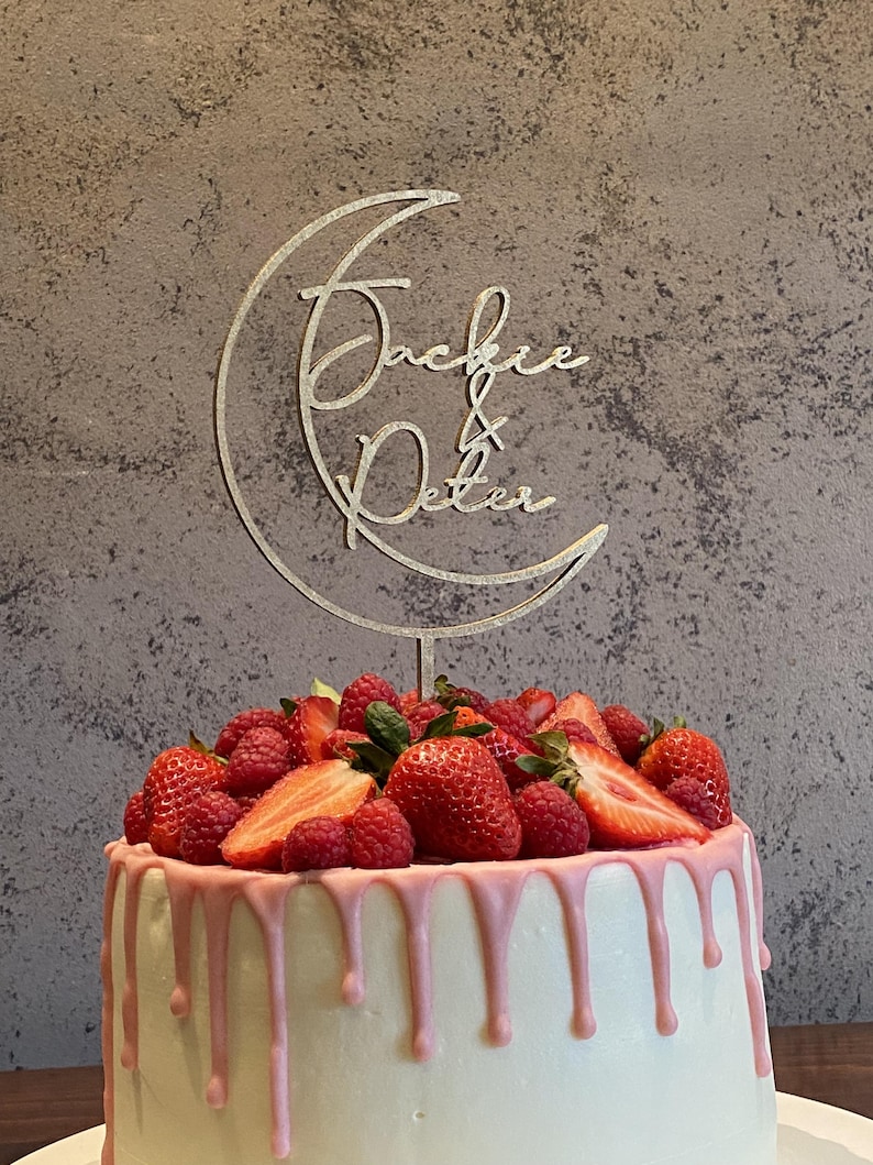 Op de afbeelding: Een witte cake met roze glazuur en verse aardbeien en frambozen. Een houten caketopper met een halve maan ontwerp en de namen "Jackie & Peter" is op de cake geplaatst.