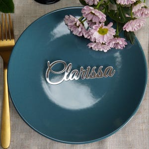 May include: A teal plate with a silver nameplate that reads "Clarissa" in cursive script. A gold fork and knife flank the plate. Pink flowers are placed on the plate. The setting is on a linen tablecloth.