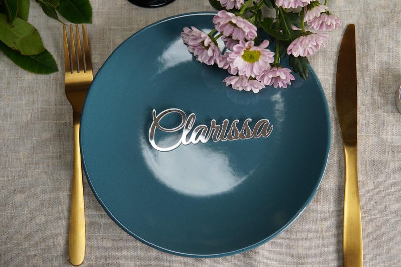 May include: A teal plate with a silver nameplate that reads "Clarissa" in cursive script. A gold fork and knife flank the plate. Pink flowers are placed on the plate. The setting is on a linen tablecloth.