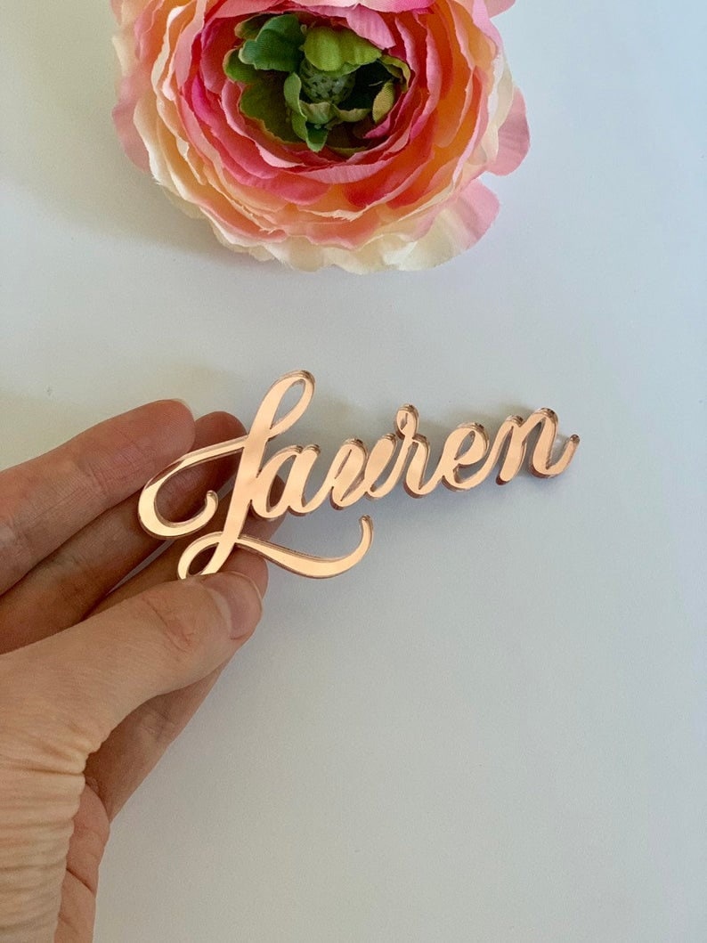 Puede incluir: Una tarjeta de nombre de acr&iacute;lico espejo de color oro rosa con el nombre "Lauren" escrito en cursiva.