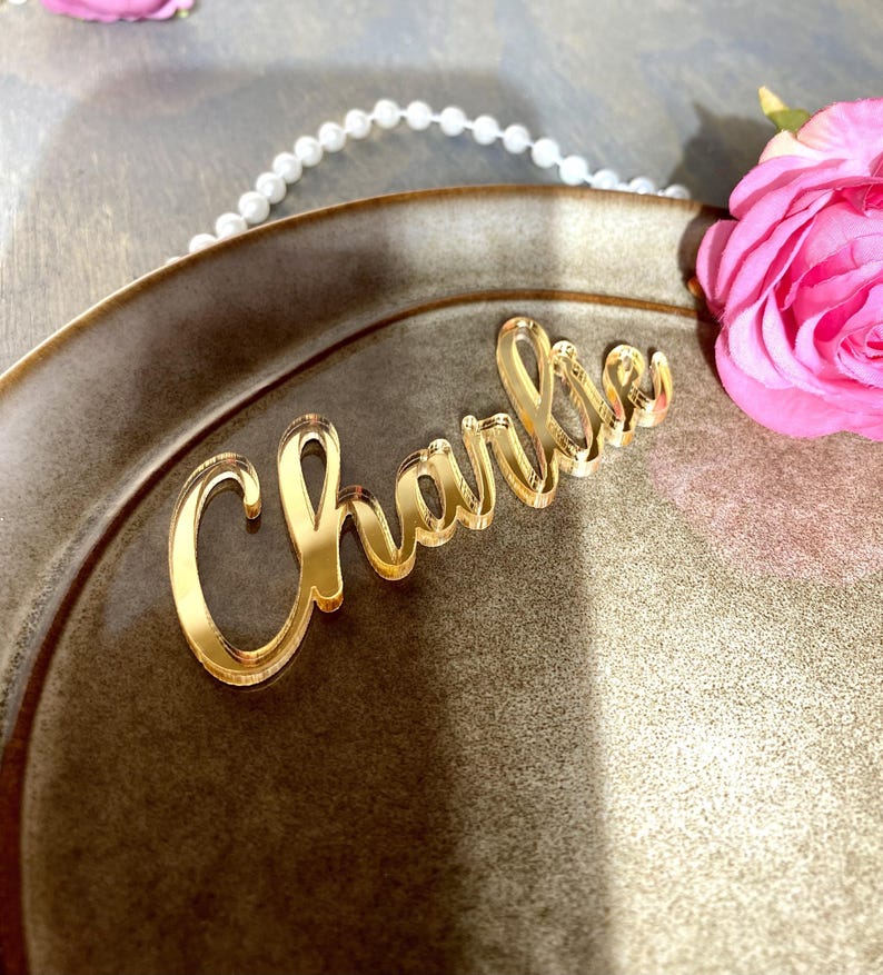 May include: A gold-colored acrylic nameplate that reads "Charlie" in a cursive font. The nameplate is placed on a brown plate, with a pink rose and a string of pearls in the background.