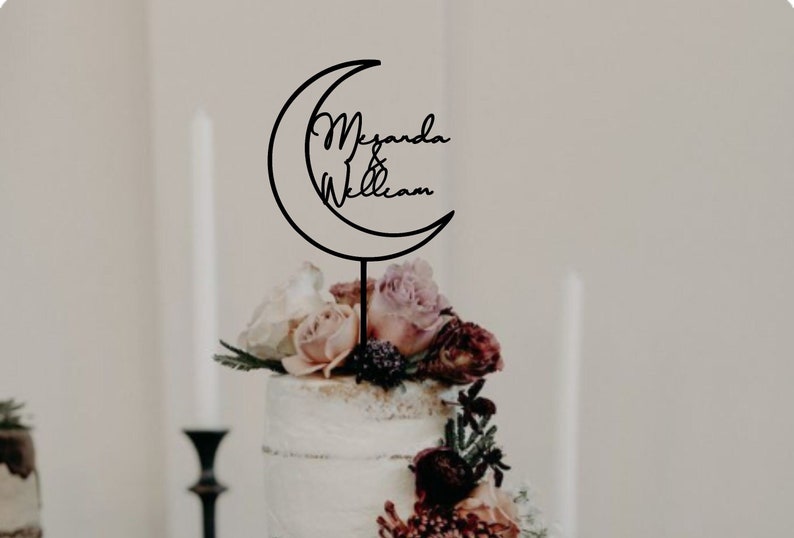Op de afbeelding: Een zwarte metalen taarttopper in de vorm van een halve maan met de namen "Meganda & William" erin geschreven. De topper is geplaatst op een witte taart die is versierd met roze en witte bloemen.