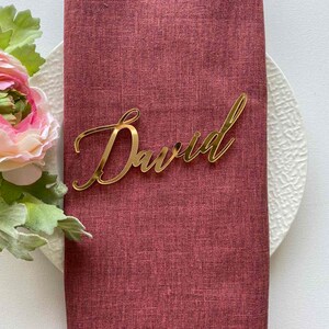 Puede incluir: Tarjeta de lugar de acr&iacute;lico dorado con el nombre "David" escrito en cursiva.