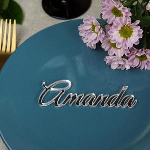 May include: A teal plate with a gold fork and a floral arrangement of pink flowers. A mirrored acrylic place card with the name "Amanda" is centered on the plate, perfect for a wedding or event.