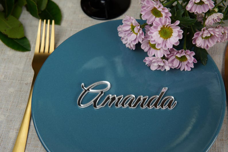May include: A teal plate with a gold fork and a floral arrangement of pink flowers. A mirrored acrylic place card with the name "Amanda" is centered on the plate, perfect for a wedding or event.