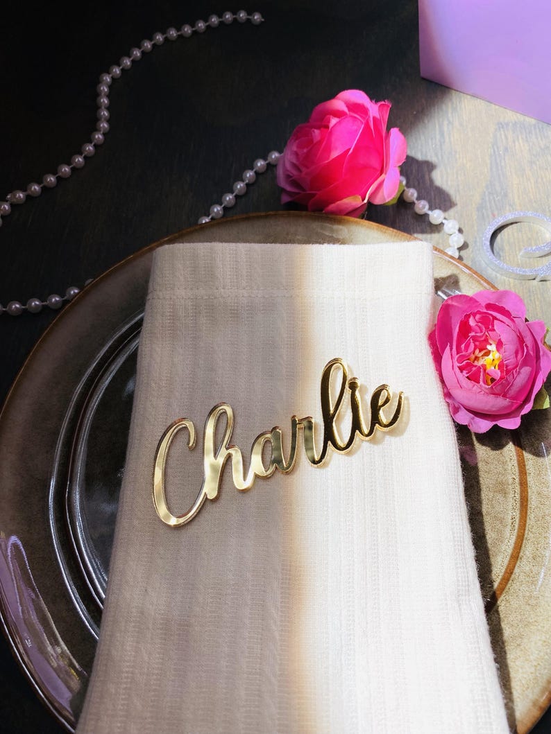 May include: A gold-colored nameplate that reads "Charlie" in cursive script rests on a white napkin. The napkin is placed on a plate, with pink roses and a string of pearls in the background. The scene suggests a wedding or special event.